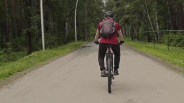 Orman yolunda bisiklet süren, sırt çantalı, koruyucu kask takan bir adamın arkasından bak.