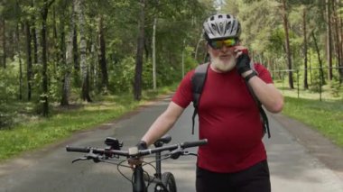 Gri sakallı, kasklı yaşlı bir adam ormanda bisikletle yürüyor ve cep telefonuyla konuşuyor.