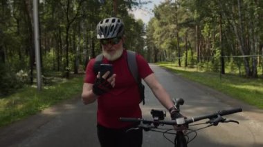Orman yolunda bisikletle gezen, yaz gezintisi sırasında cep telefonuyla selfie çeken yaşlı bisikletçi.