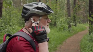 Yaşlı gri saçlı bisiklet eldivenli adam orman yolunda sürmeden önce bisiklet kaskı takıyor.