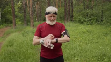 Yaşlı bir erkek koşucunun portresi. Bilekliğinde telefon var. Akıllı kaset kullanıyor ve ormanda dikilirken kameraya su şişesiyle poz veriyor.