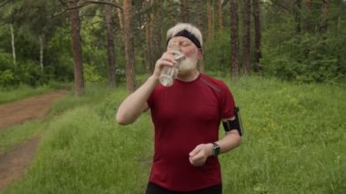 Spor giyim ve saç bandı giyen yaşlı bir adam şişeden su içiyor, fitness takip cihazını açıyor ve ormanda yolda koşuyor.