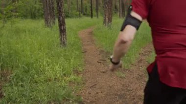 Spor giyimli yaşlı bir adamın yeşillik ve ormandaki uzun çam ağaçlarıyla çevrili yolda koşuşunun arka görüntüsü.