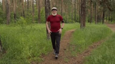 Yaşlı bir adam orman yolunda koşuyor, sonra akıllı hareket ediyor, terini siliyor ve açık hava eğitiminden sonra dinleniyor.