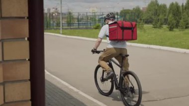 Arka planda, kuryenin başlık taktığı ve kırmızı teslimat sırt çantasıyla yerleşim alanındaki banliyö caddesinde bisiklet sürerken görüntüsü var.