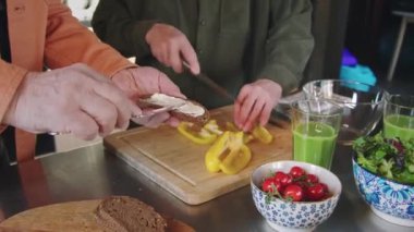 Mutfakta birlikte sağlıklı bir kahvaltı hazırlayan yaşlı çiftin yakın çekimi, kızarmış ekmeğe tereyağı süren adam, doğrama tahtasında sebze salatası için biber kesen kadın, masada yeşil smoothie.