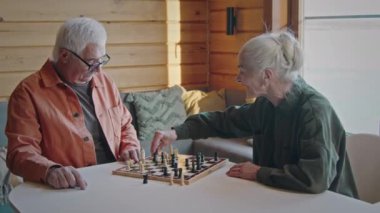 Orta yaşlı beyaz karı ve kocanın kış günü oturma odasında oturmuş satranç oynamaları, şah mat yapan, güverteye işaret eden, tezahürat yapan ve el sıkışan kadınların orta boy fotoğrafları.