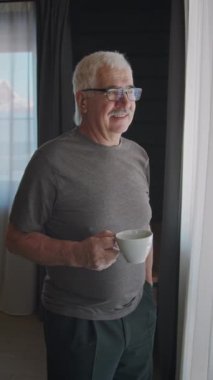 Gümüş saçlı, bıyıklı, gözlüklü, pencerenin önünde duran, sabah çayının ya da kahvenin keyfini çıkaran, kameraya bakıp gülümseyen beyaz bir adamın orta boy dikey portresi.