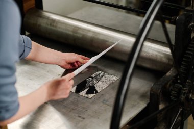 Hands of young female designer putting paper on artwork pattern while bending over printing machine with metallic tray and roller clipart