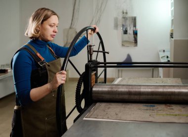 Giysi ve önlüklü genç sarışın kadın zanaatkarın atölyede ve dönen tekerlekte baskı makinesinin yanında duruşu.