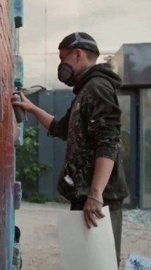 Gaz maskeli genç erkek grafiti sanatçılarının orta boy dikey pozu, lekeli kapüşonlu kapüşonlu, şehir sokaklarında ya da avluda büyük renkli duvar resimleri yapmakla meşgul.
