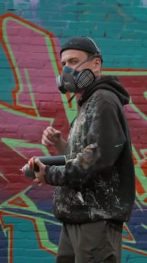 Kafkas erkek grafiticinin orta boy dikey portresi kameraya doğru dönüyor, boya şişelerini tutuyor, solunum cihazını çıkarıyor, yeni sanat çalışmalarını tamamladıktan sonra kameraya cesurca bakıyor.