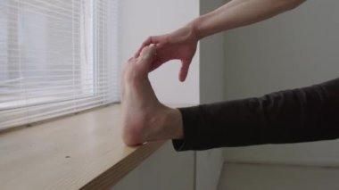 Çıplak ayaklı erkek dansçı bir bacağını pencere pervazında tutuyor ve stüdyoda provaya hazırlanırken kaslarını esnetiyor.