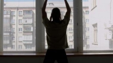 Erkek balerin siluetinin arkadan görünüşü topukluları kaldırıyor, kollarını kafasının üstüne koyuyor, sonra pencere pervazını barre olarak kullanıyor ve stüdyoda egzersiz yaparken plie çalışması yapıyor.