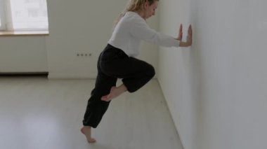 Dansçı kadın dizini göğsüne kadar çekiyor ve stüdyoda ısınırken bir bacağını duvara doğru uzatıyor.