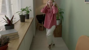 Kablosuz kulaklıklı, evde enerjik danslar yapan rahatlamış sarışın bir kız.