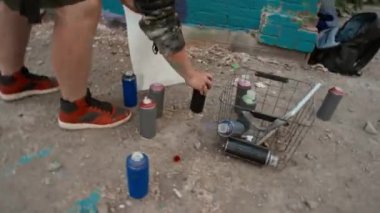 Kapüşonlu ve solunum cihazlı profesyonel erkek sokak sanatçısının el kamerasıyla çekilmiş bir fotoğrafı. Yerden bir şişe grafiti boyası seçiyor, karıştırmak için titriyor ve şehir sokaklarında yeni bir başyapıt yaratıyor.