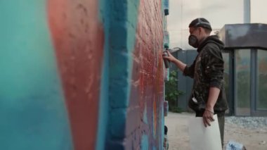 Solunum cihazlı, şapkalı, kazaklı genç bir erkek duvar ressamı arka bahçedeki büyük renkli duvarlara son rötuşları yapıyor.