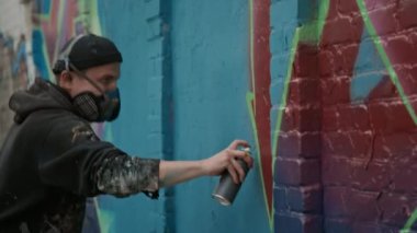 Kimyasal koruma solunum cihazı, şapka ve kapüşonlu genç beyaz adamın yeşil sprey boyayla tuğla duvardaki kırmızı-mor graffiti etiketinin etrafını çizerken orta boy fotoğrafı.