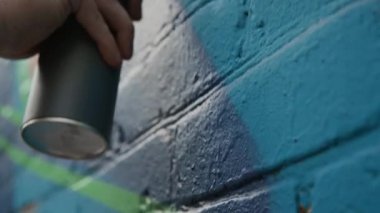 Şehir sokaklarında mavi tuğlalı duvarda grafiti üzerinde çalışırken yeşil sprey boyayla çizim yapan tanınmayan şehir sanatçılarının yakın çekimi.