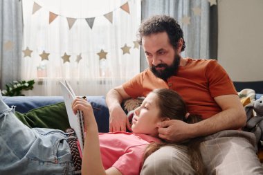 Baba, dekoratif unsurlar ve pencerelerden süzülen doğal ışıkla rahat oturma odasında otururken kanepeye yaslanıp kızına kitap okuyor.