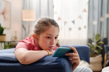 Genç kız, yıldız temalı dekor, bitkiler ve yumuşak ışıklandırmayla dekore edilmiş oturma odasında koltukta dinlenirken akıllı telefon kullanıyor.