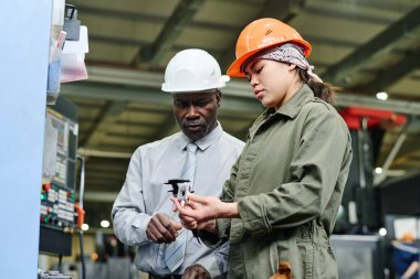 İki fabrika işçisi, bir kıdemli adam ve bir genç kadın, endüstriyel bir ortamda makinelerin onarımını tartışıyorlar, ikisi de güvenlik kaskı takıyorlar ve görevlerine odaklanıyorlar.