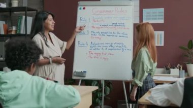 Orta boy kızıl saçlı beyaz saçlı bir kız okuldaki İngilizce dersinde dil bilgisi kurallarıyla beyaz tahtada duruyor İspanyol bayan öğretmen sorular soruyor ve öğrenci cevap veriyor.