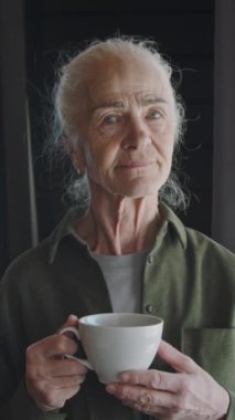 Yüzünde kırışıklıklar olan 70 yaşındaki beyaz bir kadının orta boy dikey portresi, çörekte uzun gri saçlar, sabah kahvesinin keyfini çıkarıyor, kameraya bakıyor, gülümsüyor.