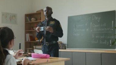 Siyahi bir polis memurunun kariyer günü boyunca okul çocuklarıyla konuşurken, polis kimliği ve telsizini gösterirken, öğrencilere mesleğini ve görevlerini anlatırken orta boy fotoğrafı.