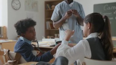 Orta boy Afro-Amerikalı bir erkek ve Orta Doğulu bir kız okuldaki kariyer dersi sırasında steteskopla oynarken tanınmayan siyahi bir doktor yakınlarda onu izliyor.