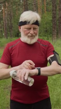 Spor giyim ve saç bandı takan yaşlı bir adamın dikey portresi. Akıllı kasetteki fitness uygulamasını kullanıyor ve ormanda kameraya poz veriyor.