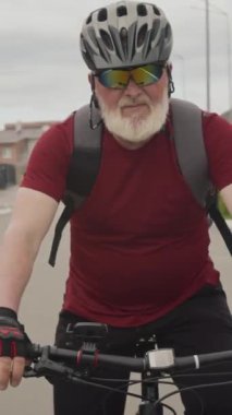 Miğferli ve güneş gözlüklü yaşlı adamın dikey görüntüsü mahalledeki yolda kameraya doğru dönüyor.