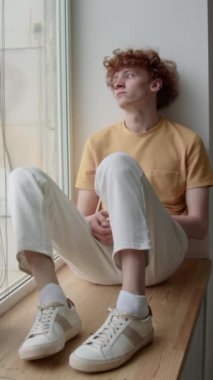 Dikey çekimde, günlük giysiler ve spor ayakkabılar giymiş beyaz bir adam evdeki ahşap pencere pervazında oturmuş, derin düşünceler içinde, geçmişteki olaylardan pişmanlık duyarak sokağa bakıyor.