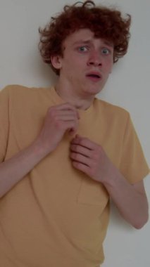 Hardal tişörtlü genç, korkmuş beyaz adamın dikey portresi kameraya bakıyor ve yüzünü elleriyle kapatıyor, korkmuş ve kapalı alanda korunmasız hissediyor.