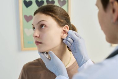 Eldiven giyerken genç hastaların kulaklarını muayene eden profesyonel bir doktor. Arka plandaki kalpler rahatlatıcı bir atmosfer yaratıyor.