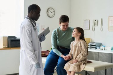Afrikalı Amerikalı doktor klinik ortamında oturan çocuk hasta ve ebeveyniyle ilgileniyor. Görünür tıbbi ekipman ve duvara monte edilmiş saat klinik ortamı güçlendirir