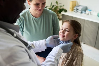 Afro-Amerikalı doktor beyaz önlük giyip sağlık kliniğinde genç bir kızı muayene ediyor anne odaklanmış ifadeleri izliyor ve tıbbi çevreyi önemsiyor.