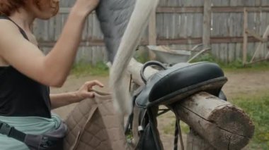 Genç at binicisinin, yaklaşan sürüş için hazırlanan ahşap korkuluklara dikkatlice at eyeri ve ped yerleştirdiği orta ölçekli bir fotoğraf.