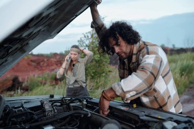 Siyah dalgalı saçlı genç adam kamyonetin kaputunu eğip motoru kız arkadaşıyla karşılaştırıyor ve tamir servisini arıyor.