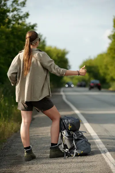 stock image Rear view of young woman in casual attire standing by highway in front of her backpack and catching pickup truck or other vehicle