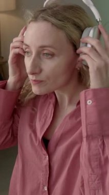 Dikey çekimde genç sarışın kadın evde duruyor, kulaklık takıyor ve en sevdiği müzikle dans ediyor.