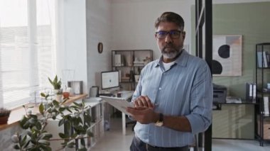 Gözlüklü, çizgili mavi gömlekli, dijital tabletle modern ofise poz veren ve kameraya bakan olgun sakallı Hintli şirket yöneticisinin orta boy portresi