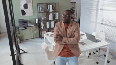 Dolly, modern ofiste kollarını kavuşturup kameraya bakan, gözlüklü, şık giyinen siyahi iş adamının portresi.