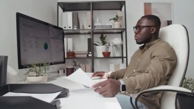 Gözlüklü Afrikalı Amerikalı erkek ofis çalışanının belgeye bakıp verileri bilgisayara girerken, mali rapor doldururken yan görüntüsü.