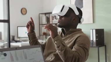 Genç Afro-Amerikan erkek şirket yöneticisinin modern artırılmış gerçeklik kulaklığıyla ofis toplantısında el kol hareketi yaparken göğüs göğse çekilmesi.