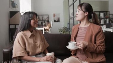 Orta boy İspanyol ve Asyalı iş kadınlarının gözlüklü ve şık gündelik elbiseli kahve fincanlarıyla kahverengi deri koltukta otururken ve ofis toplantısından sonra sohbet ederken.