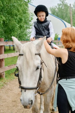 Siyah kasklı ve yelekli çocuk çiftlikte beyaz ata biniyor, çitli yolda çocuğa rehberlik ederken dizginleri elinde tutan kadın, arka planda yeşil ağaçlar.