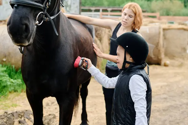 stock image Young boy wearing helmet brushing horse under supervision of woman on farm. Both engaging in bonding activity, horse standing calmly while being groomed