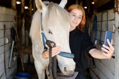 Beyaz atlı beyaz bir kadının ahırda selfie çektiği beyaz bir kadın. Ahşap duvarları ve sağlam bir iç yapısı olan insan ve hayvan arasındaki bağı gösteriyor.